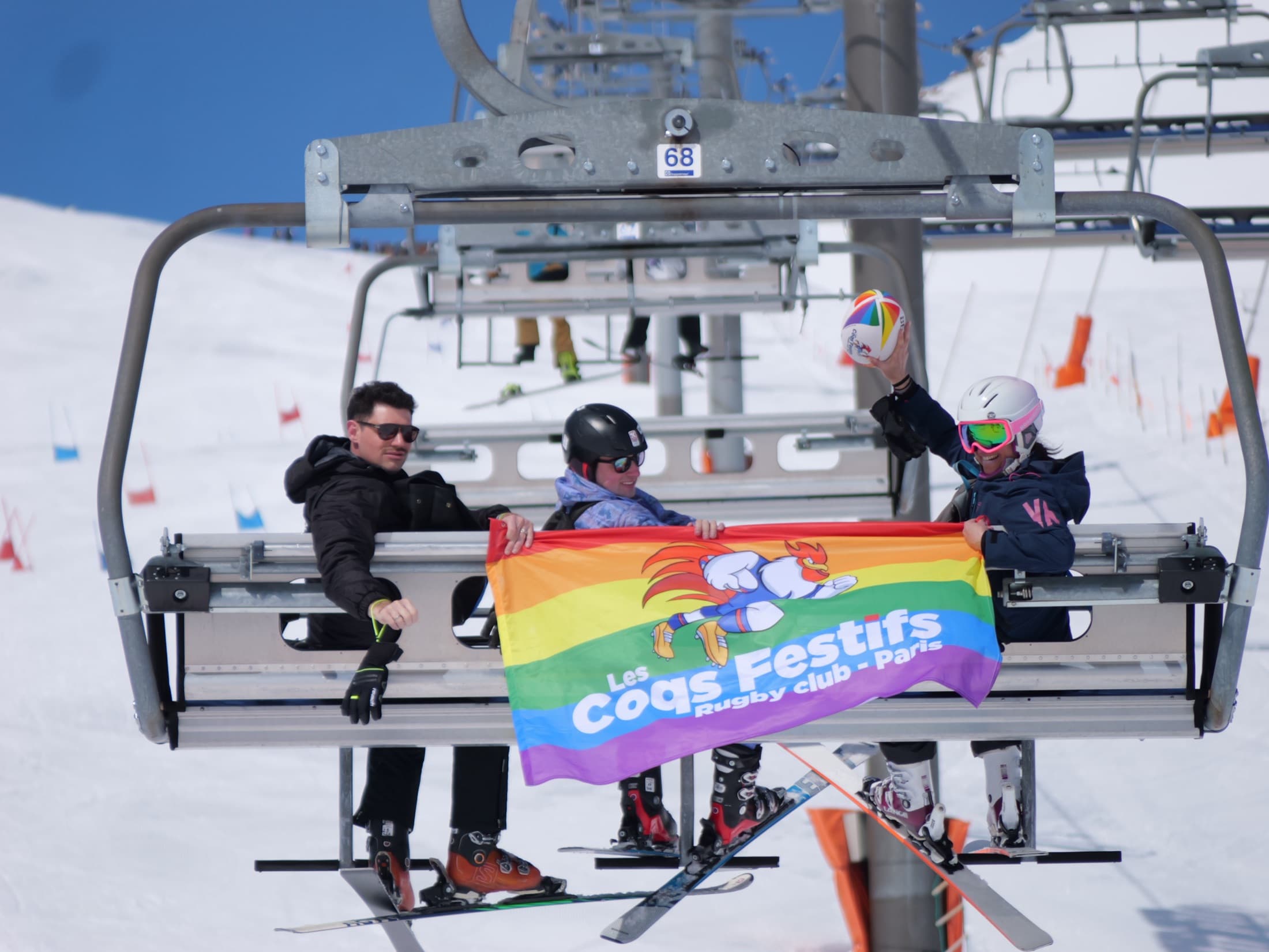 Les Coqs Festifs sur un télésiège avec leur drapeau et un ballon arc-en-ciel
