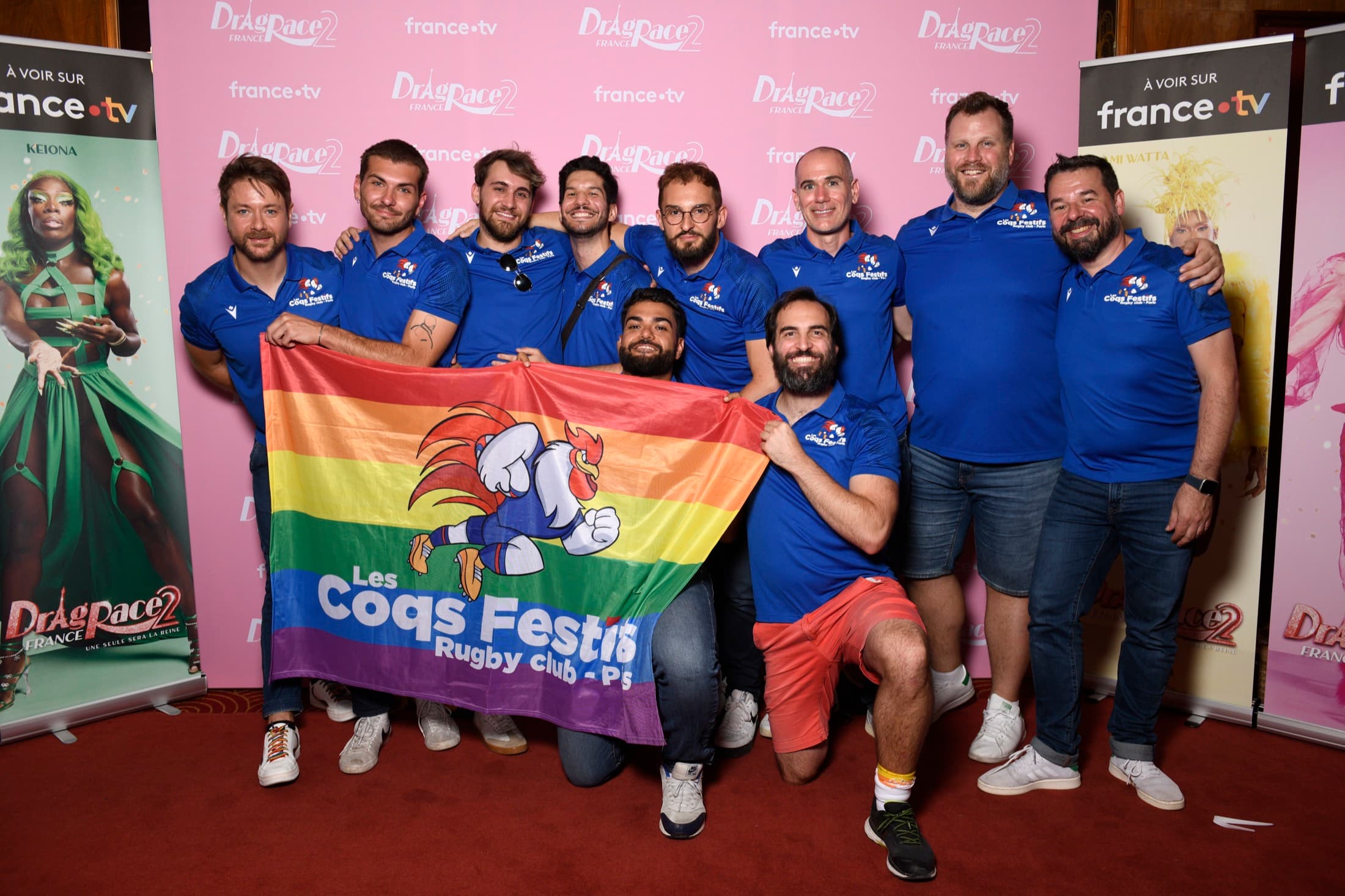 L’équipe des Coqs Festifs réunie autour du drapeau du club devant des affiches Drag Race France