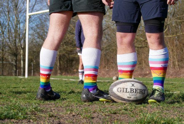 Chaussettes de rugby arc-en-ciel des Coqs Festifs sur le terrain, à côté d'un ballon Gilbert
